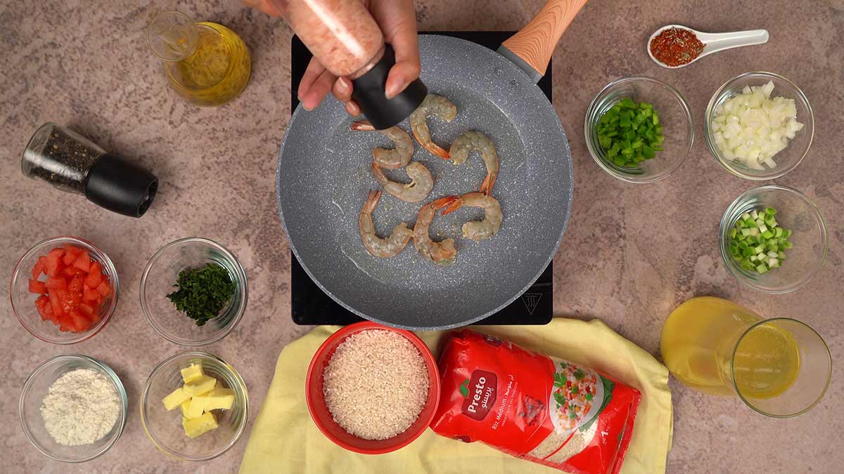 des crevettes assaisonnées dans une poêle entourée d'ingrédients frais comme le riz, les légumes, les épices et le bouillon.