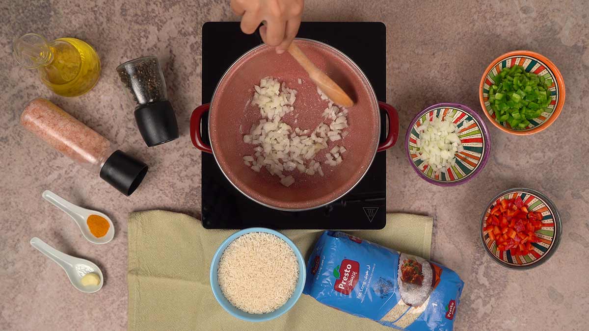 La préparation du riz au curry et poivrons, où les oignons sont en train de revenir dans une casserole, entourés d'ingrédients comme les poivrons rouges et verts, du riz, du curcuma, de l'ail en poudre, et des assaisonnements.