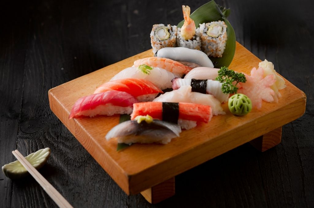 Un plateau de sushis variés, incluant des nigiris au saumon, au thon, à la crevette et au maquereau, accompagnés de makis tempura, de wasabi, et de décorations en forme de fleurs, soigneusement disposés sur un support en bois pour une présentation élégante.