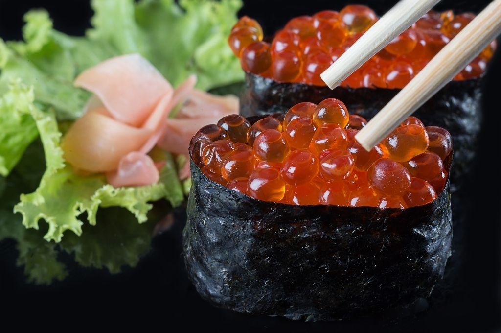 Des gunkans garnis d'œufs de saumon brillants, enveloppés de nori, avec une garniture de gingembre mariné et de feuilles de salade en arrière-plan, offrant une présentation raffinée et appétissante.