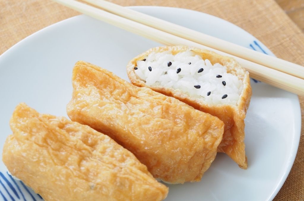 Inarizushi des poches de tofu frit remplies de riz vinaigré, agrémentées de graines de sésame noir, joliment disposées sur une assiette blanche avec des baguettes.