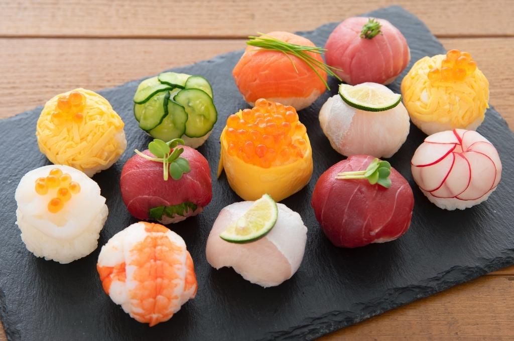 Une belle sélection de temari sushi, des boulettes de riz garnies de poissons variés, crevettes, tranches de citron vert, radis, concombres, œufs et œufs de poisson, soigneusement disposées sur une ardoise noire pour une présentation élégante et colorée.