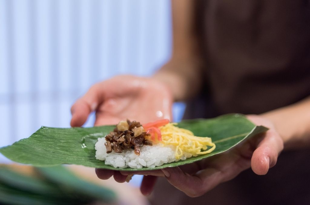 Préparation de sushi sur une feuille de bambou, contenant du riz vinaigré garni de morceaux de viande cuisinée, d'omelette râpée et de gingembre mariné, tenue dans les mains pour une présentation artisanale et authentique.