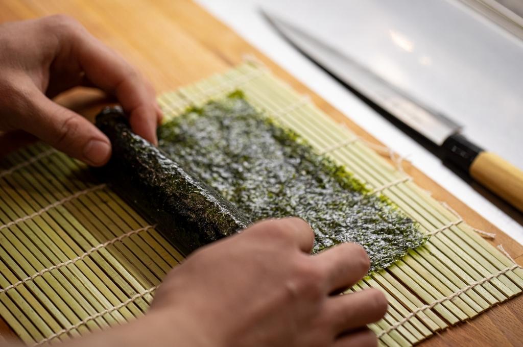 La préparation de sushi, où une personne roule une feuille d’algue nori sur une natte en bambou, un couteau traditionnel étant posé à côté.