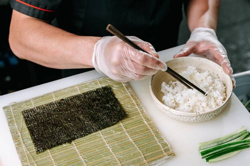 Un homme en train de préparer du sushi, en utilisant des baguettes pour prendre du riz dans un bol et le disposer sur une feuille de nori posée sur une natte en bambou.