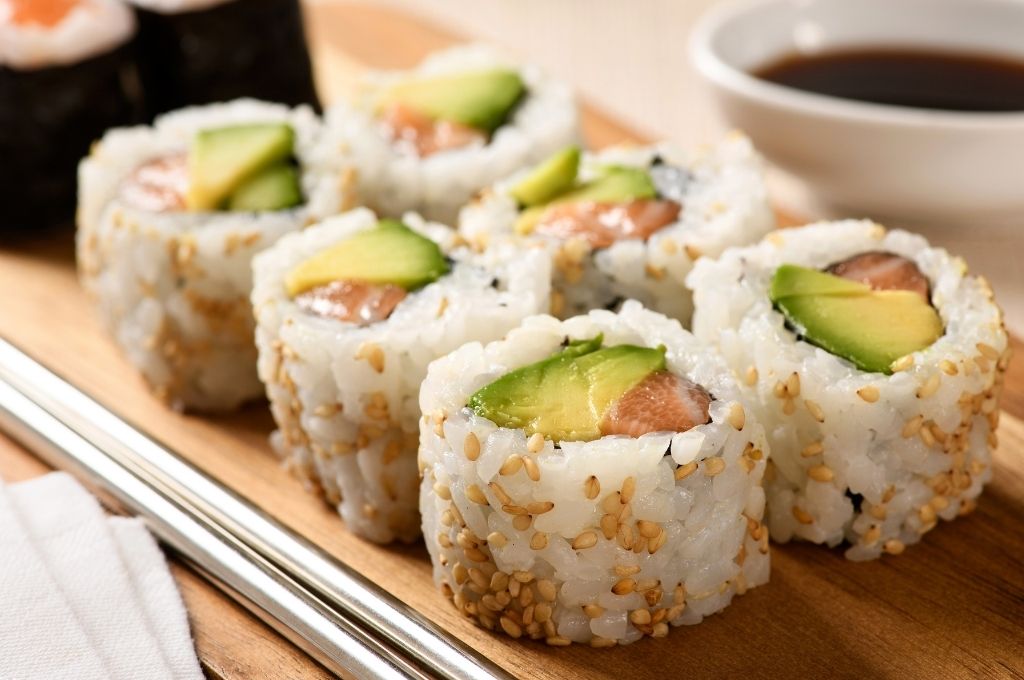 Sushis California un des types of sushi rolls garnis d'avocat et de saumon, enrobés de graines de sésame, disposés sur un plateau en bois, accompagnés d'une coupelle de sauce soja.