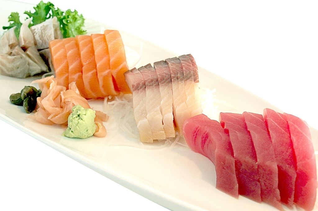 Assortiment de sashimi disposé sur une assiette blanche, incluant du thon, du saumon, du maquereau et d'autres poissons frais, accompagné de gingembre mariné, de wasabi et de quelques garnitures vertes pour une présentation élégante et épurée.