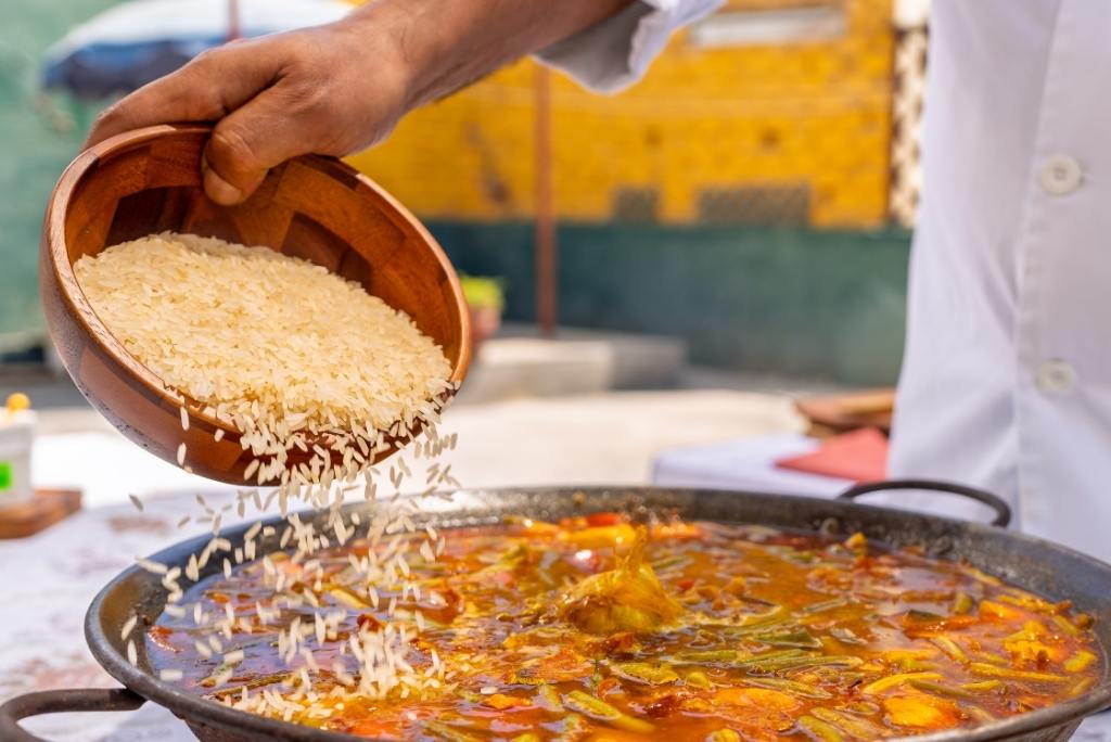 أيدٍ تسكب الأرز من وعاء خشبي في مقلاة بايلا كبيرة أثناء التحضير.