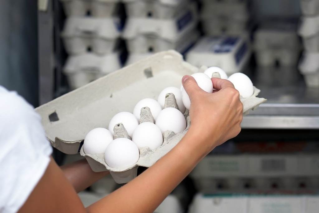 Gros plan sur des mains qui manipulent des œufs blancs dans une boîte en carton, avec d'autres boîtes en arrière-plan.