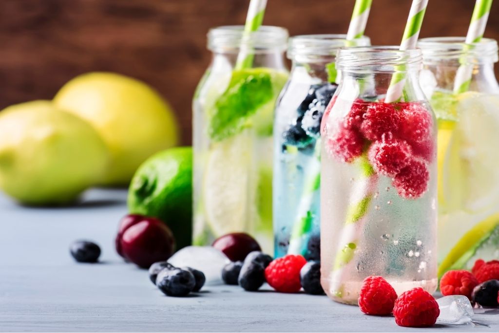Assortiment de boissons nutritives et rafraîchissantes dans des bouteilles en verre, garnies de fruits frais comme des framboises, des myrtilles, des citrons verts et des citrons, parfaites pour l'hydratation.