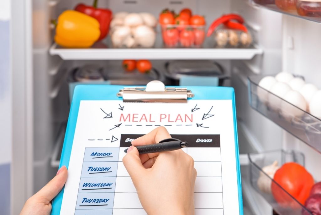 Une personne utilisant un stylo pour remplir un plan de repas hebdomadaire sur un presse-papiers devant un réfrigérateur ouvert rempli de légumes frais, symbolisant l'organisation et la planification des repas sains.