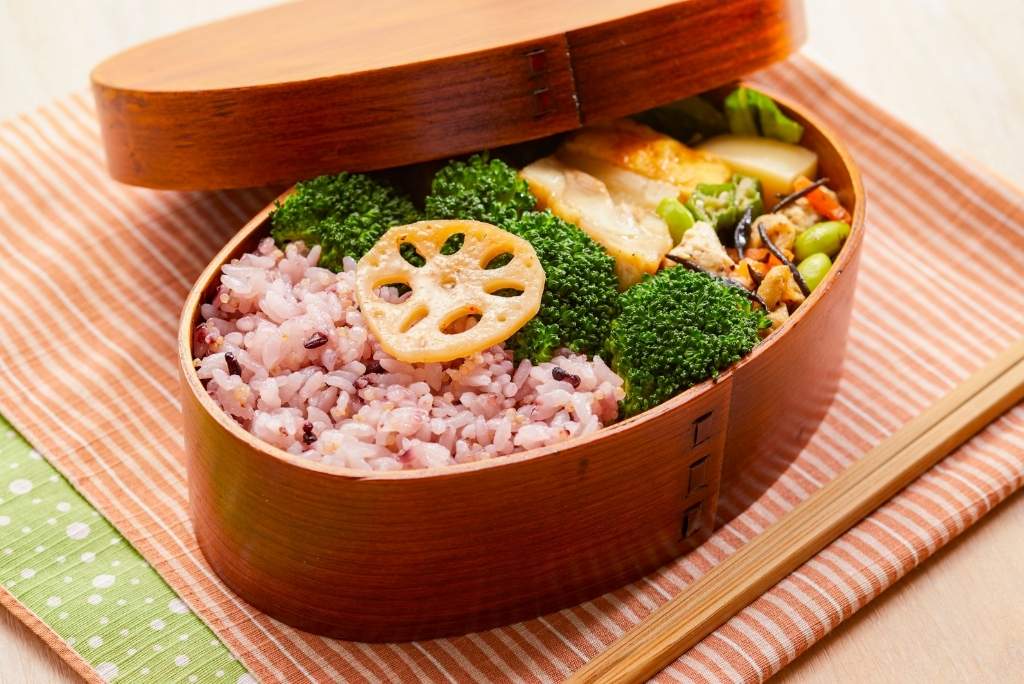 Une boîte à bento traditionnelle en bois ouverte, remplie d'un plat équilibré de riz complet et d'une variété de légumes colorés comme le brocoli et une tranche de lotus, prête pour un repas sain.