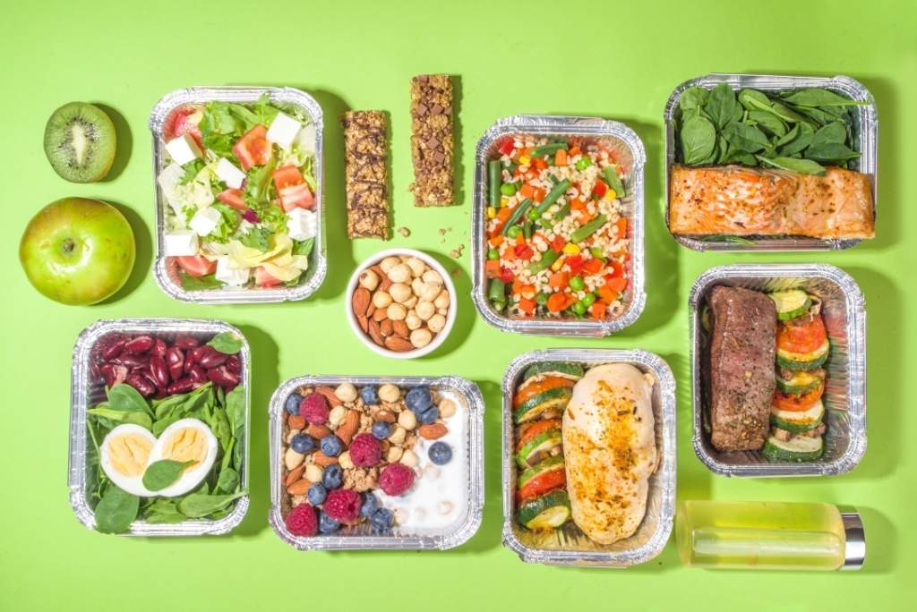 Vue de dessus de plusieurs contenants de repas préparés et emballés pour la semaine, incluant du poisson, des légumes, des salades, des légumineuses et des noix, idéaux pour les repas à emporter au travail.