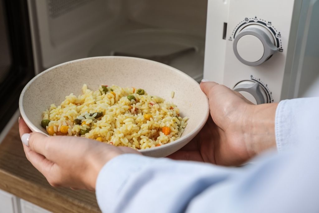 استفيدوا من الميكروويف يد شخص يضع وعاء أرز بالخضروات داخل فرن ميكروويف أبيض.
