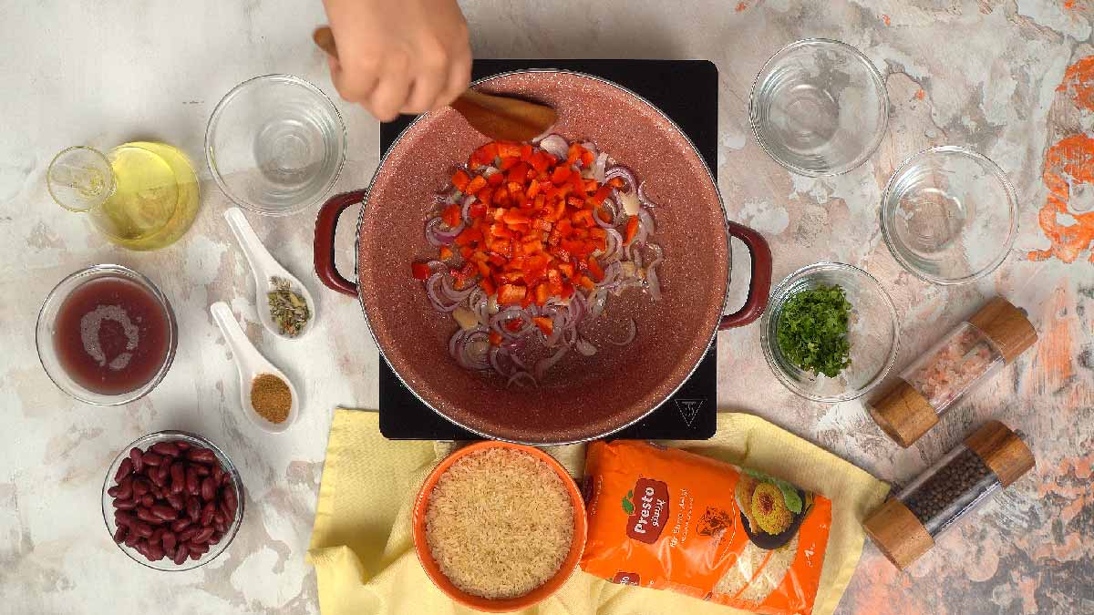 Une personne mélange des dés de poivrons rouges avec des oignons rouges hachés dans une casserole.