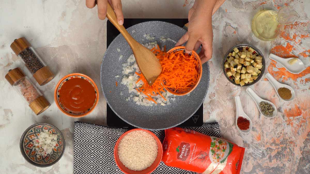 Une personne ajoute des carottes râpées aux oignons qui cuisent dans la poêle. 