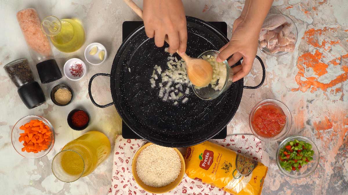 Deux mains versent des oignons hachés dans une poêle remplie d'huile au centre d'une table avec d'autres ingrédients de cuisine.
