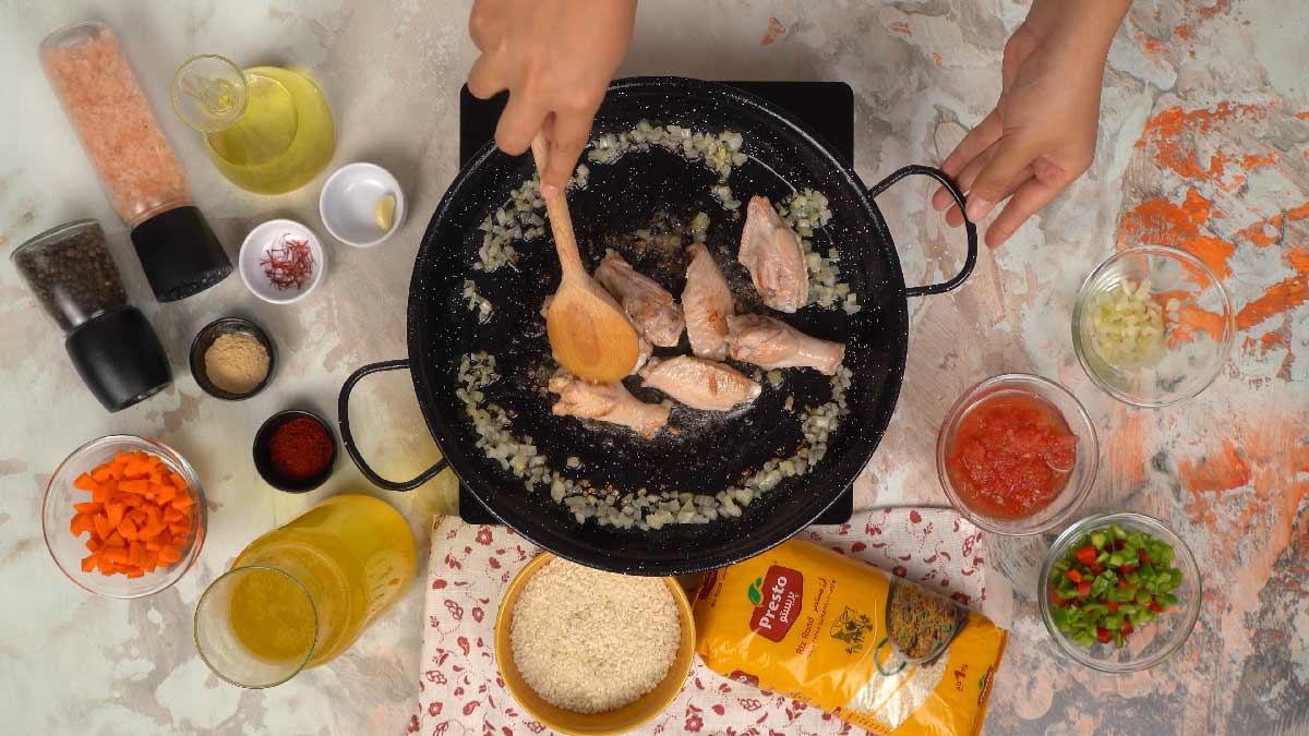 Des mains utilisent une cuillère en bois pour mélanger des morceaux de poulet et des oignons hachés dans une poêle.