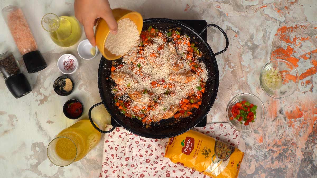 Une main verse du riz rond Presto dans bol jaune directement dans la poêle sur les légumes et le poulet.
