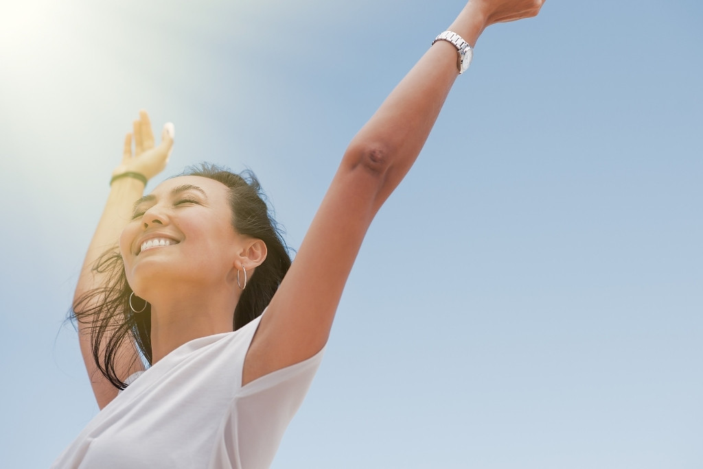 Une femme souriante, les bras levés vers un ciel bleu et ensoleillé, symbolisant la joie et l'exposition au soleil.