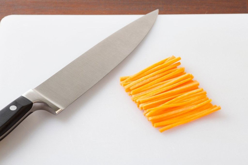 Un couteau de chef posé à côté d'une pile de carottes coupées en julienne sur une planche blanche.