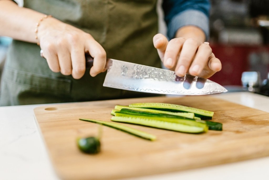 Des mains coupent des concombres en longues tranches sur une planche à découper en bois.