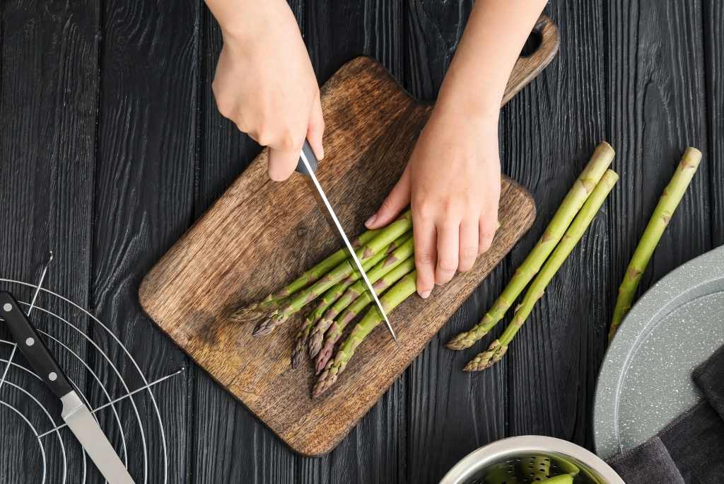 Vue d'en haut de mains coupant des asperges vertes sur une planche à découper en bois sombre. 