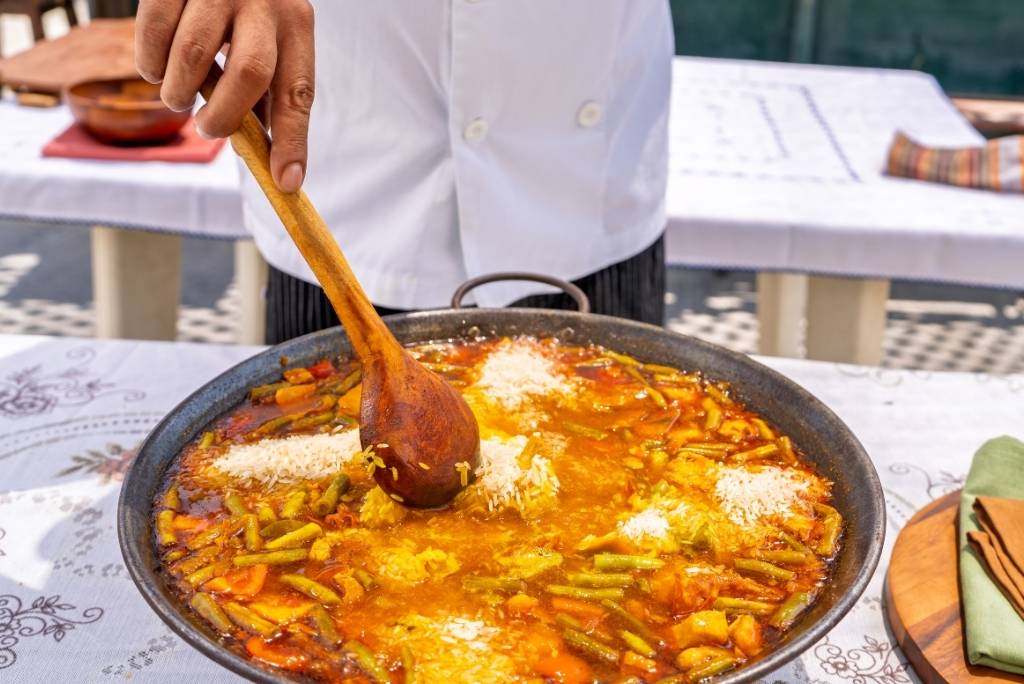طاهٍ يستخدم ملعقة خشبية كبيرة لتحريك الأرز في مقلاة باييلا أثناء التحضير.