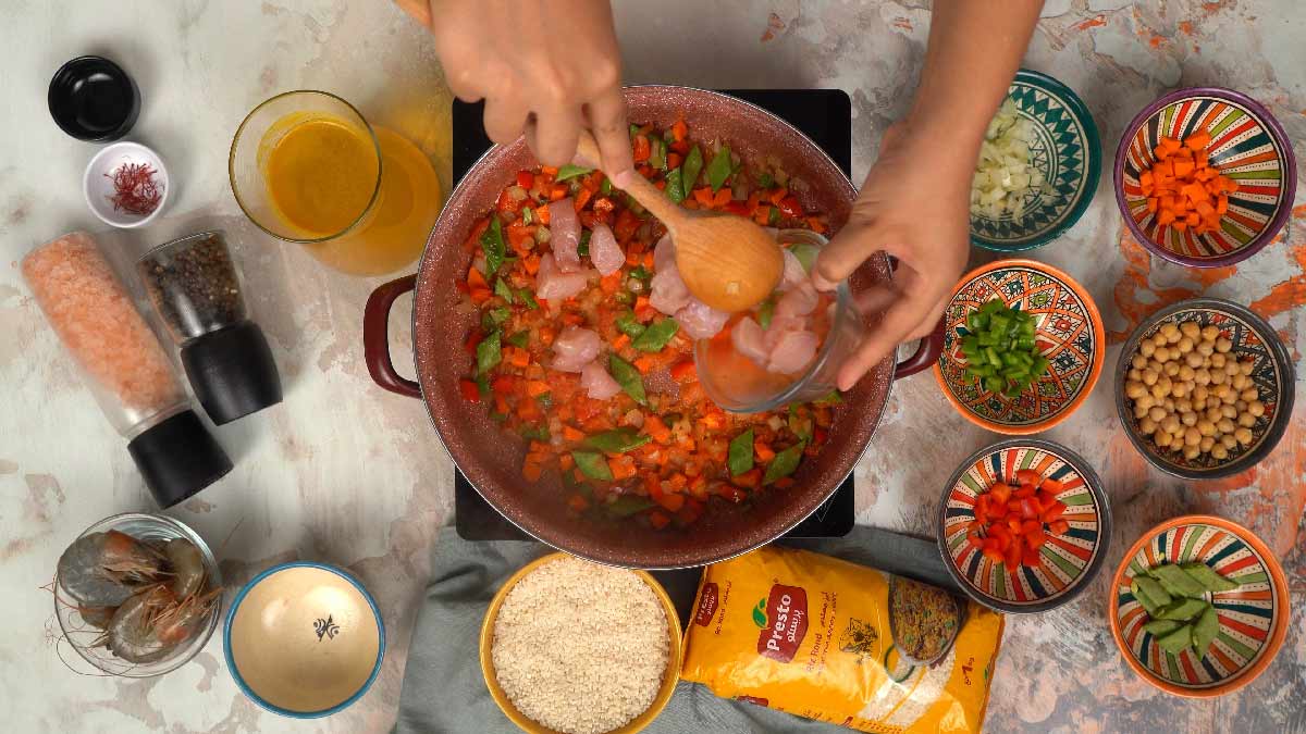 Une main utilise une cuillère en bois pour mélanger des morceaux de poulet et des légumes dans la poêle.
