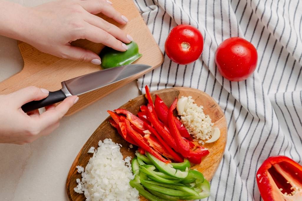 صورة من الأعلى لأيدٍ تقطع الفلفل الأخضر والأحمر على لوح تقطيع.