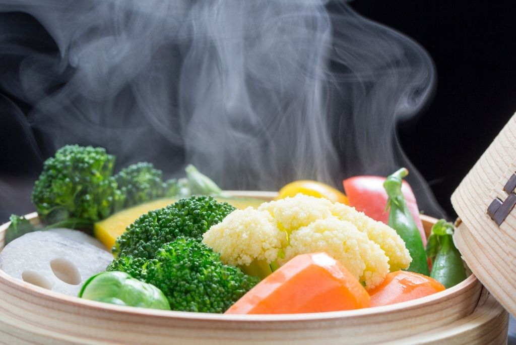 تشكيلة من الخضروات الملونة المطهية على البخار داخل سلة خشبية، يتصاعد منها بخار كثيف.