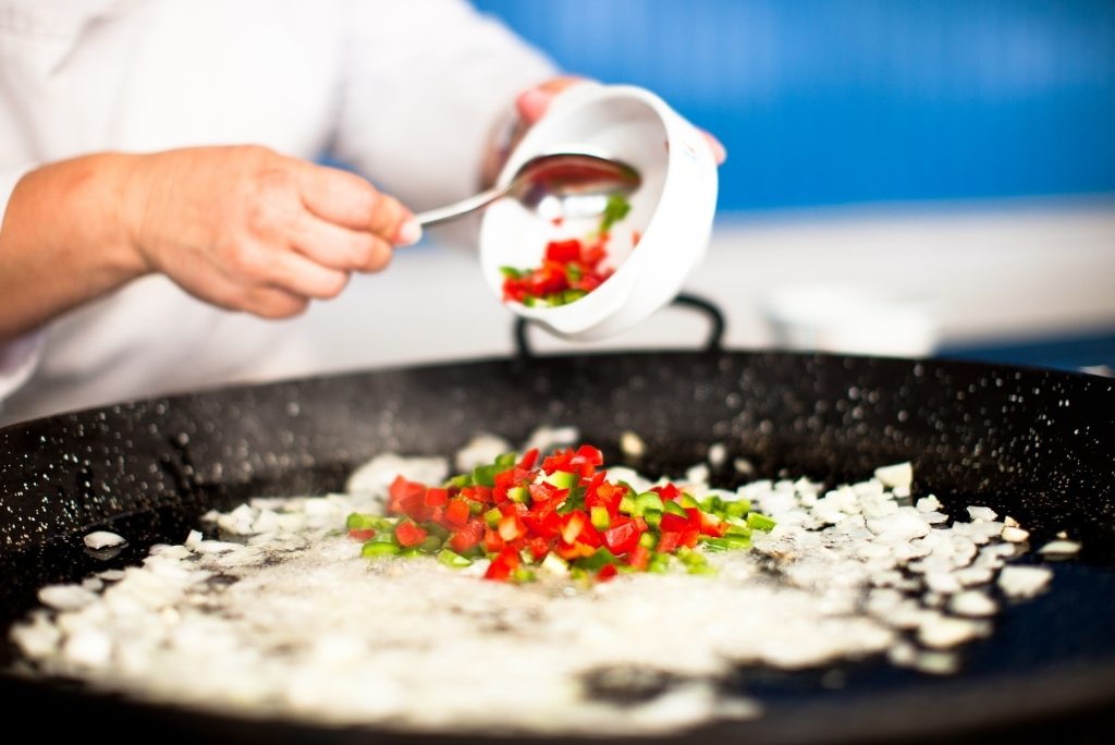 المكوّنات الأساسية لتحضير سوفريتو مثالي طاهٍ يضيف الفلفل المقطّع إلى مقلاة بايلا كبيرة تحتوي على بصل مقلى.
