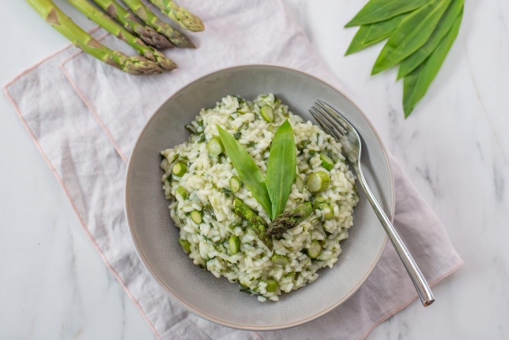 Un plat de risotto aux asperges décoré de feuilles vertes et d'une fourchette, posé sur une surface en marbre.