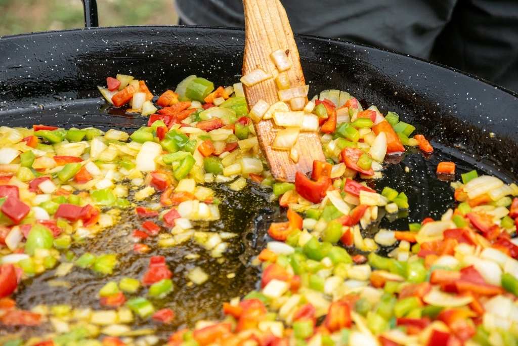 أصول وتعريف السوفريتو لقطة قريبة لبصل وفلفل يُطهى في مقلاة كبيرة مع ملعقة خشبية.