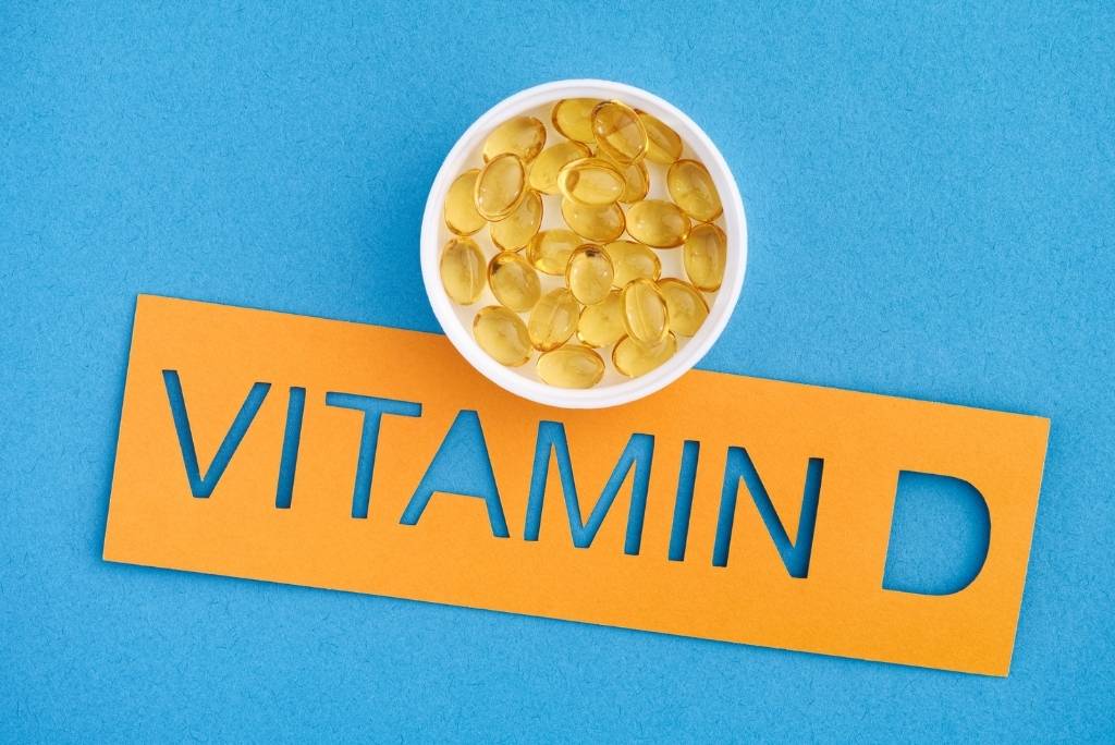 Un petit bol de gélules de vitamine D sur un fond bleu, avec des lettres découpées en papier orange épelant « VITAMIN D ».