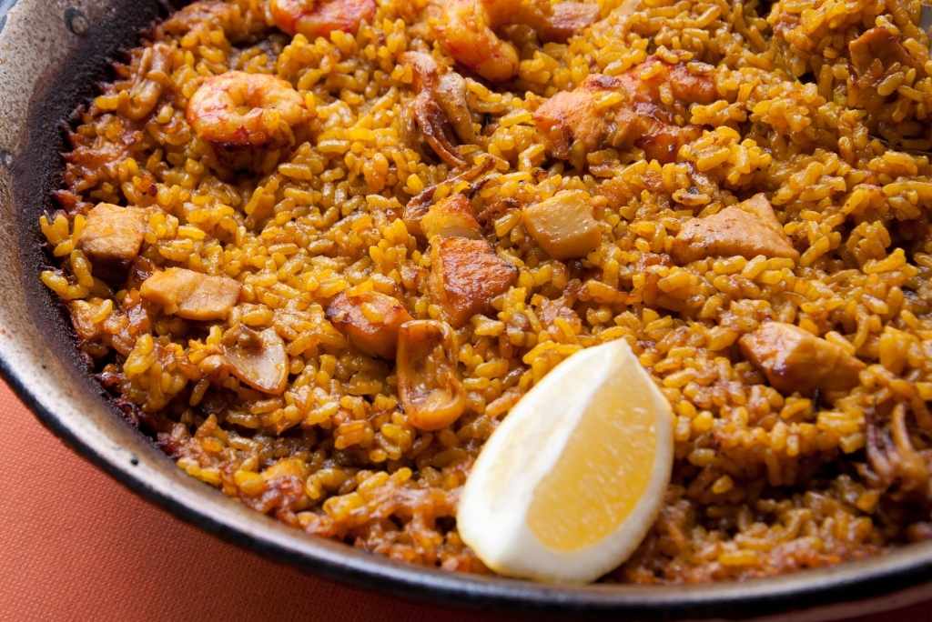 لقطة مقرّبة لباييلا مشكلة مطهية، مزينة بأرز أصفر بالزعفران، وروبيان، وحلقات كالامار، وقطع لحم، وقطعة ليمون طازجة.