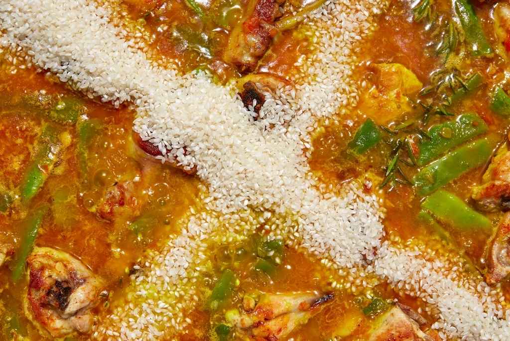 منظر علوي يوضح المرحلة الحاسمة لإضافة الأرز إلى المرق الذي يغلي برفق، حيث يُوزع على شكل صليب لضمان التوازن الصحيح بين الأرز والسائل.