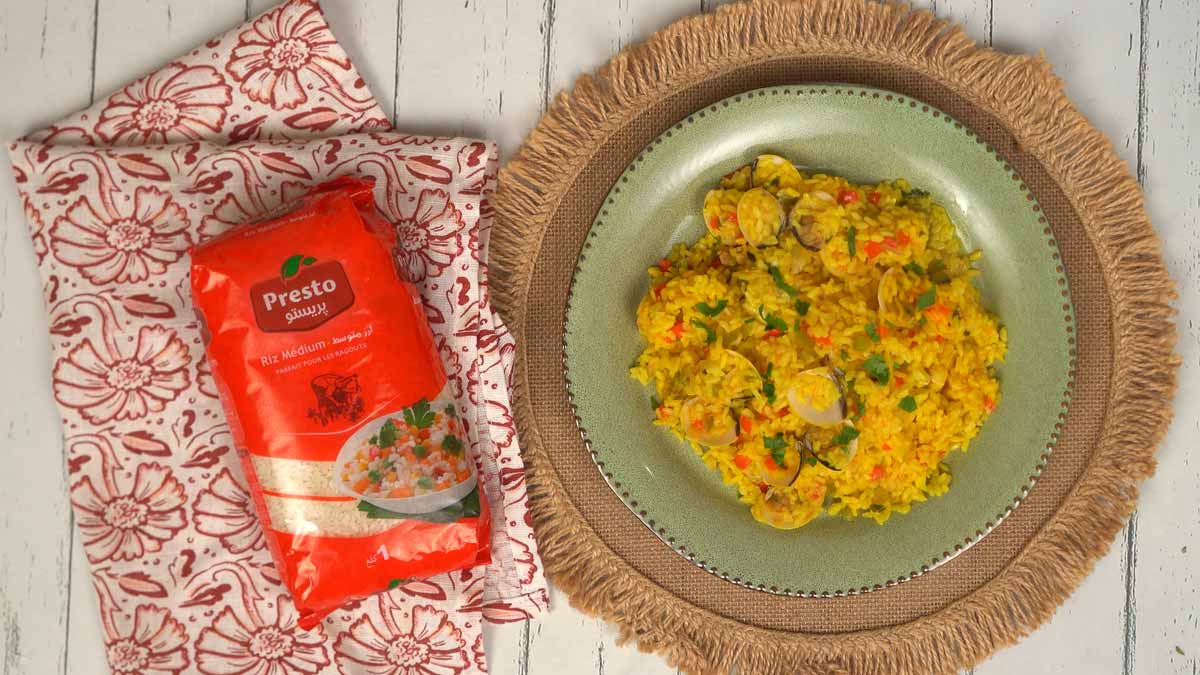 Le plat est servi dans une assiette verte, garni de persil frais, offrant un riz jaune safrané parsemé de palourdes et de petits légumes.