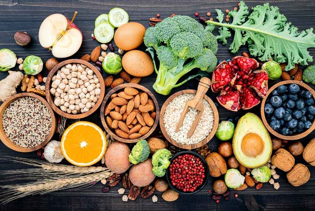صورة علوية ملونة ومتناسقة لأطعمة صحية (البروكلي، الأفوكادو، الرمان، اللوز، البيض، التوت والأرز) موضوعة على طاولة خشبية سوداء.