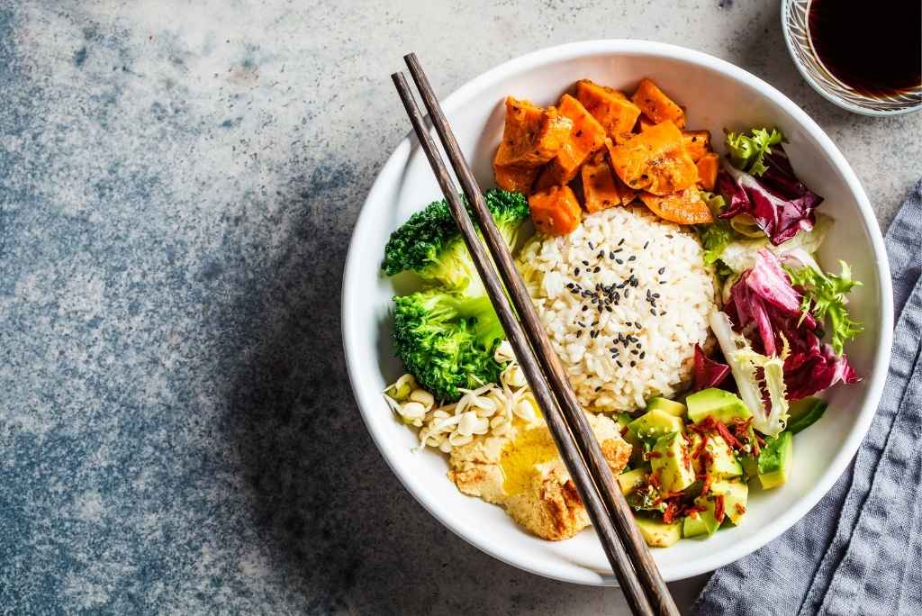 Un bol composé de riz complet entouré de brocolis, de patates douces rôties, d'avocat, de houmous et de chou rouge.